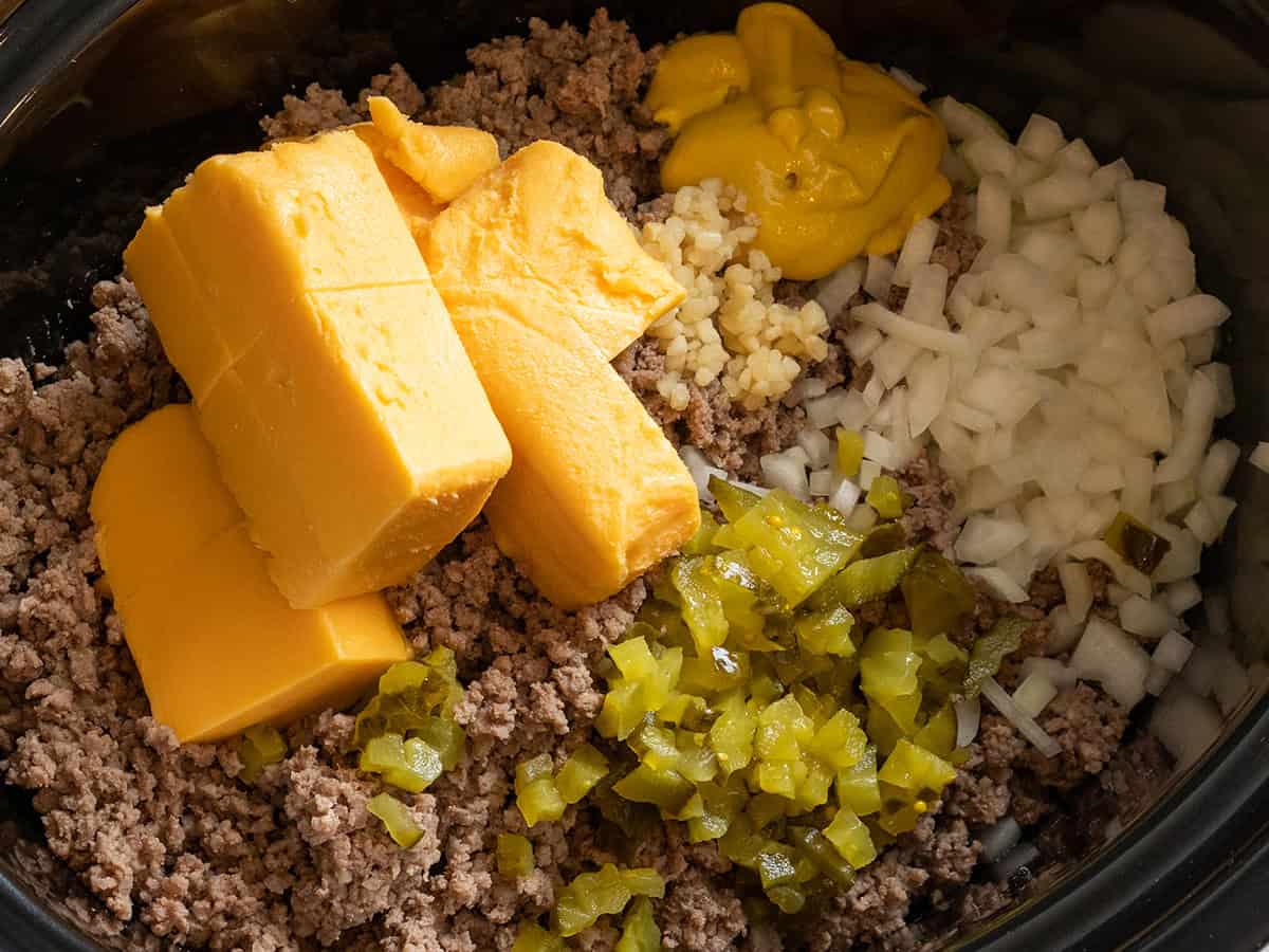 Oignon haché et cornichons, ail, moutarde, fromage Velveeta et bœuf haché doré sont dans un crockpot, prêts à être mélangés et cuits pour préparer des cheeseburgers au crockpot !