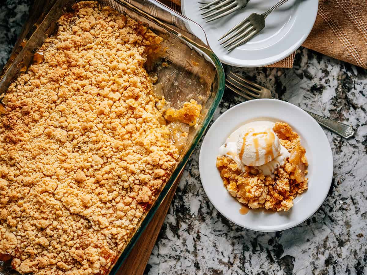 Plan de dessus d'une portion de gâteau aux pommes dans une assiette avec de la glace à la vanille sur le côté, le plat restant de gâteau aux pommes, et une pile d'assiettes vides et de fourchettes dans le coin.