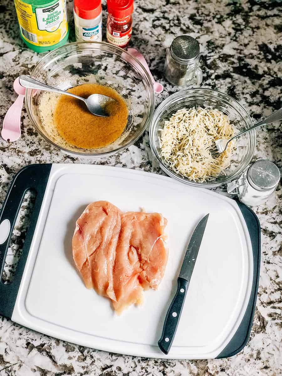 Un blanc de poulet cru ouvert en papillon sur une planche à découper, un bol de marinade à l'ail, et un bol de garniture au parmesan.