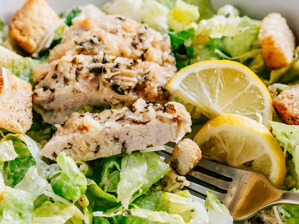 Gros plan sur un bol de laitue romaine fraîche recouvert de vinaigrette César, garni de croûtons croustillants, d'une pincée de parmesan et d'un morceau tendre et juteux de poulet au parmesan à l'ail cuit.