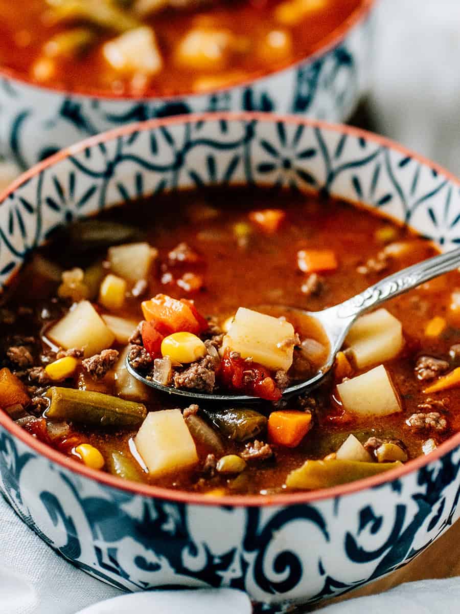 Une cuillère pleine de soupe de hamburger contenant du maïs, des haricots verts, des carottes, des pommes de terre, des oignons et d'autres assaisonnements.