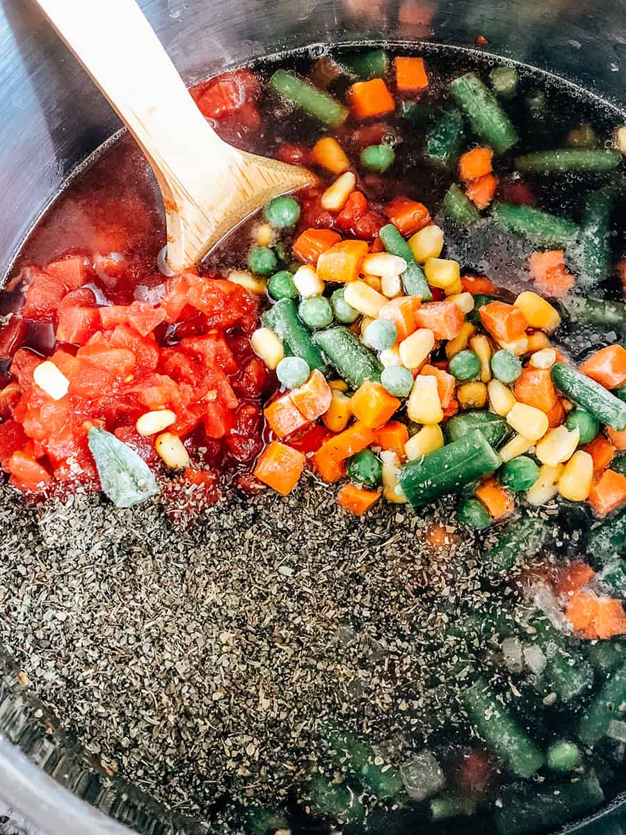 Tomates en dés petites, bouillon de bœuf, légumes congelés et assaisonnements ajoutés dans la casserole avec l'ail et les oignons.