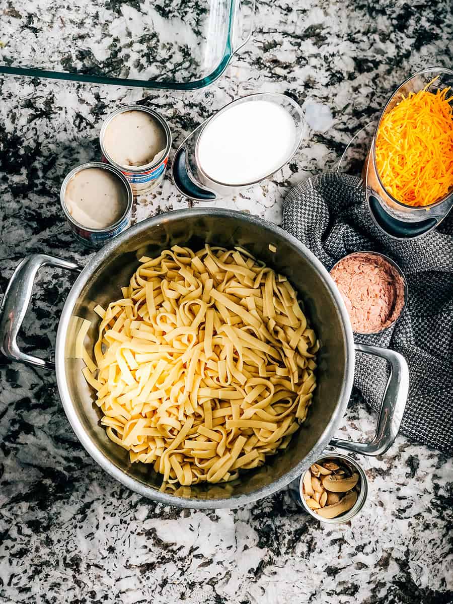 Une casserole de nouilles aux œufs cuites entourée de boîtes ouvertes des autres ingrédients, lait et fromage cheddar.