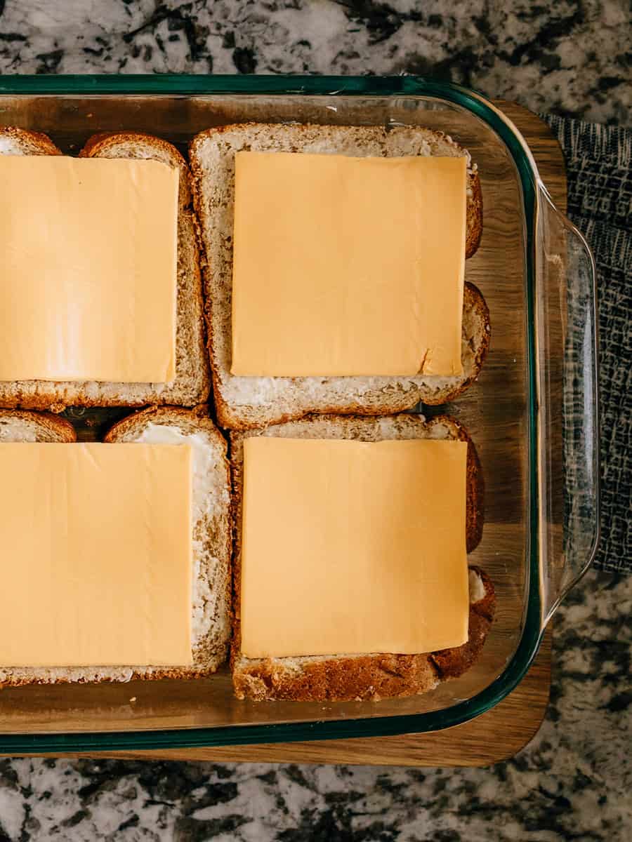 Image de pain beurré (côté beurré vers le haut) avec une tranche de fromage au-dessus dans un plat de cuisson. Six de ces pains constituent la couche inférieure de cette casserole.