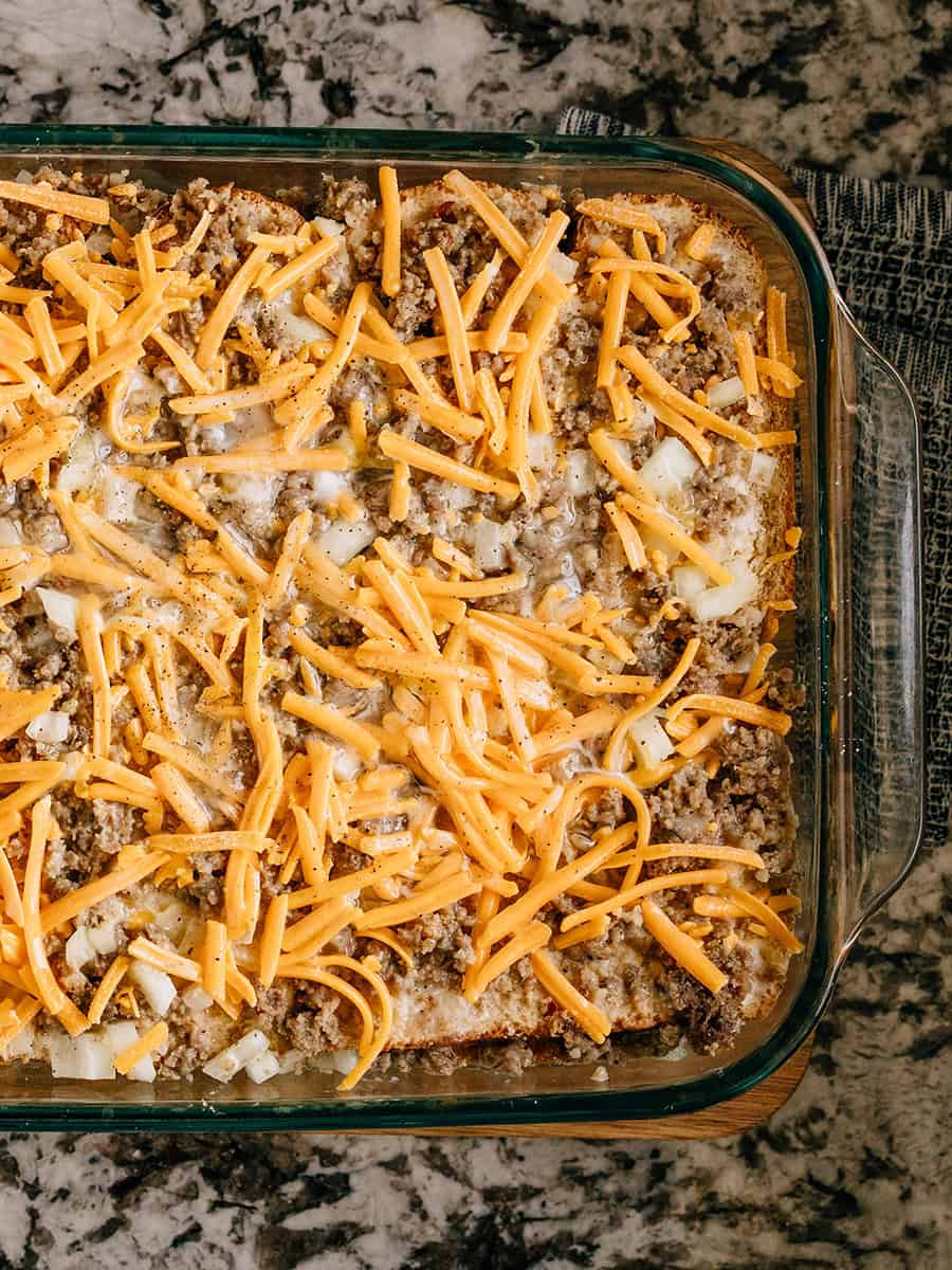 Répétez le pain beurré et saupoudrez de plus de saucisse hachée et d'oignons. Ajoutez du fromage cheddar râpé et versez le mélange d'œufs sur le tout.