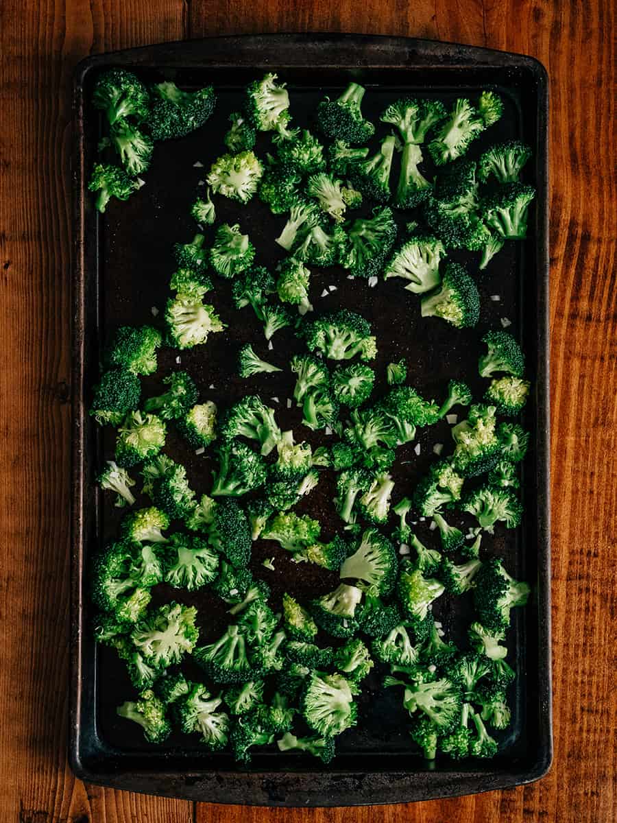 Des fleurons de brocoli frais découpés à la même taille et mélangés avec de l'huile, du jus de citron, de l'ail émincé et du sel, étalés sur une plaque de cuisson prêts à être rôtis.