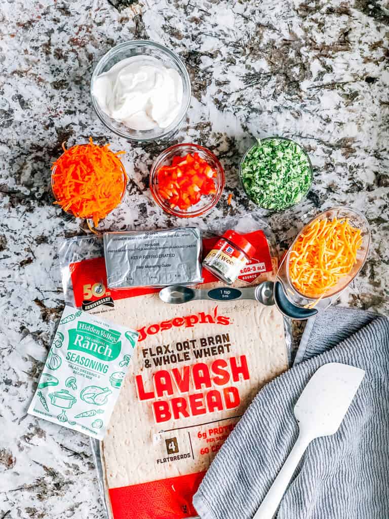 Les ingrédients pour préparer des rouleaux de légumes : pain lavash, assaisonnement ranch, fromage à la crème, aneth, poivron haché, brocoli haché et carottes et fromage cheddar râpé.