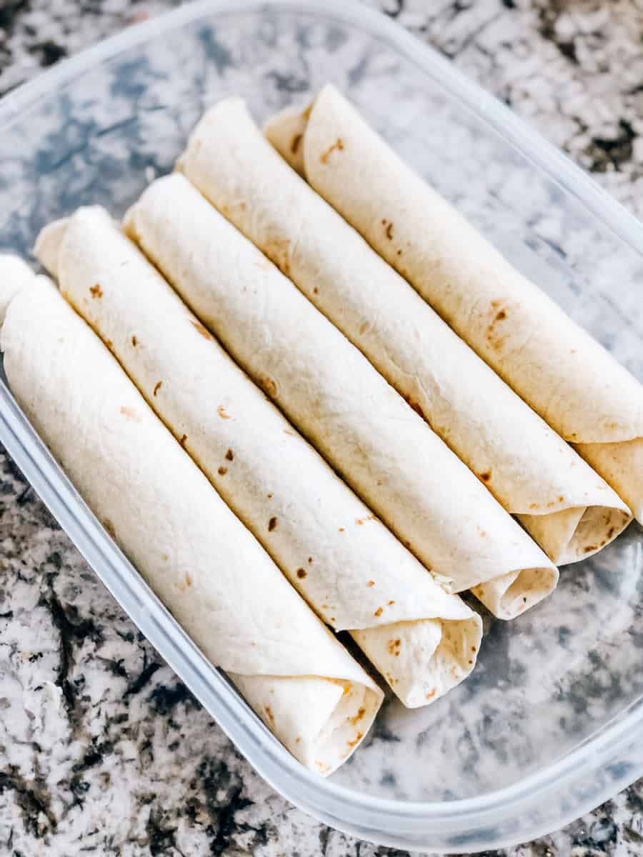 Des tortillas surdimensionnées enroulées dans un grand conteneur Tupperware prêtes à être réfrigérées.