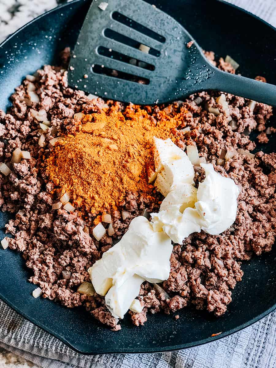 Viande hachée cuite avec oignon haché dans une poêle avec assaisonnement à taco et fromage à tartiner ramolli sur le dessus, prête à être mélangée.