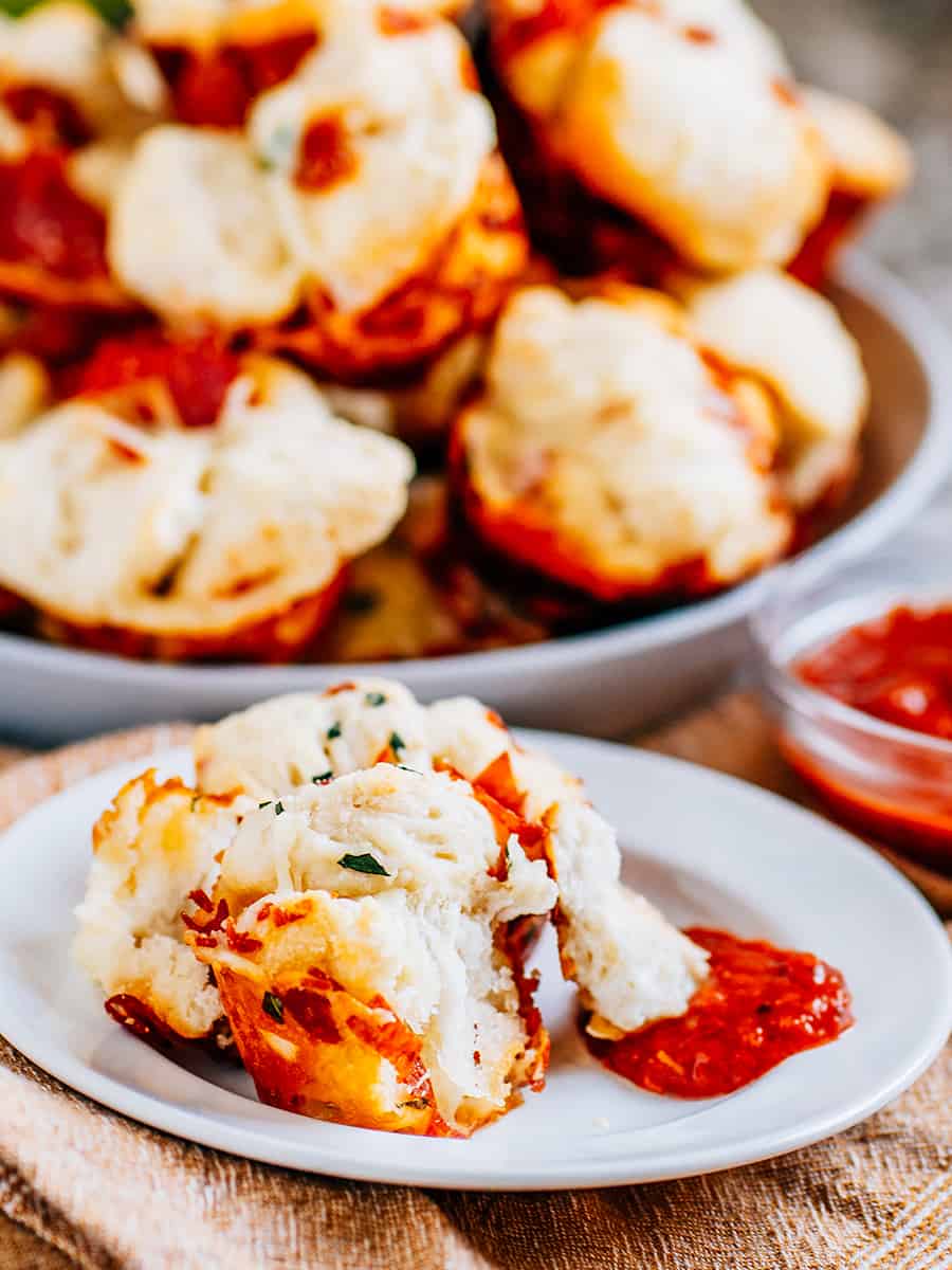 Muffins à pizza à déchirer sur une assiette avec un côté de marinara et une pile de muffins en arrière-plan.