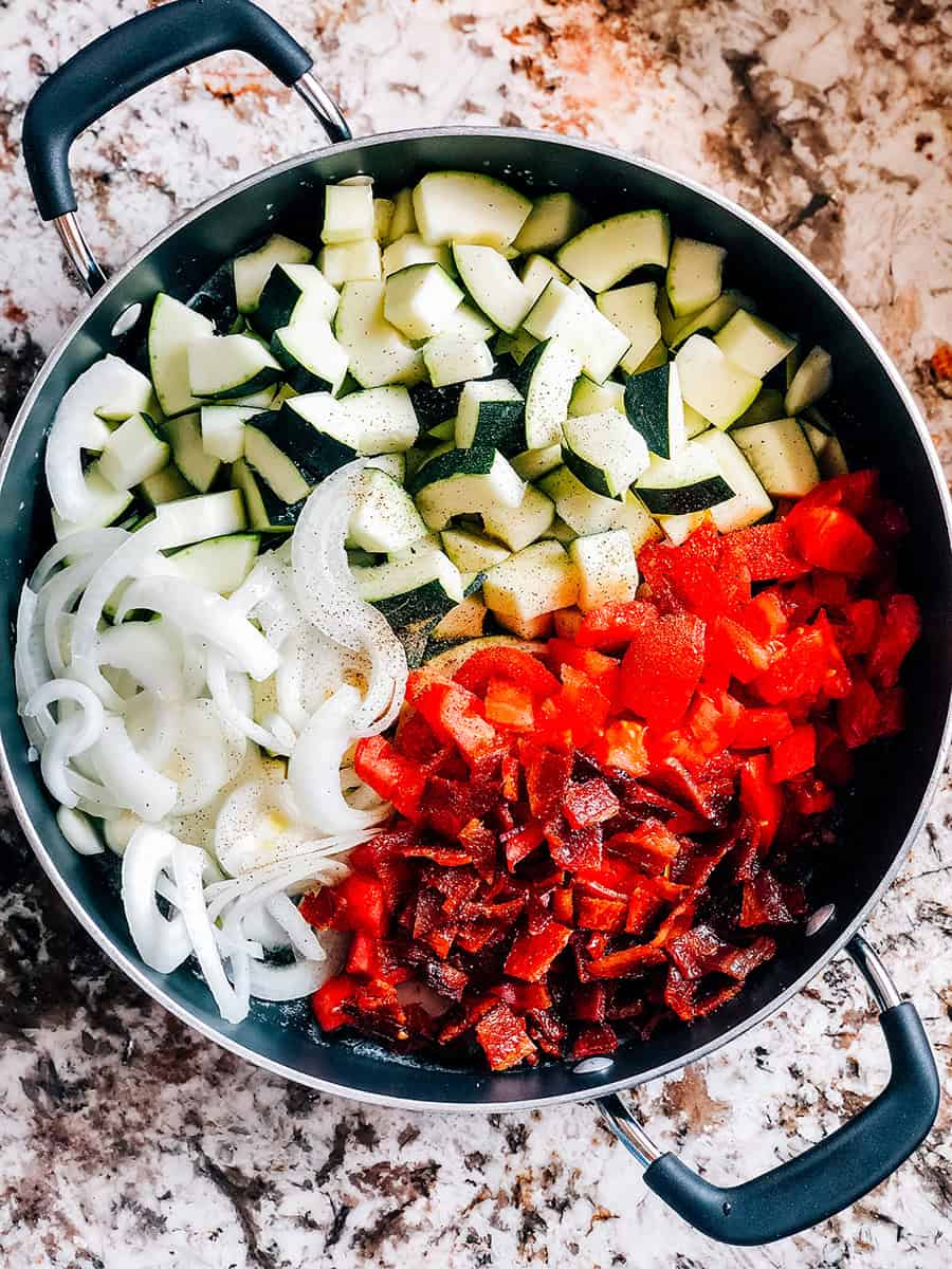 Une poêle pleine de tomates en dés et de courgettes, de bacon émietté et d'oignons tranchés assaisonnés de sel et de poivre.