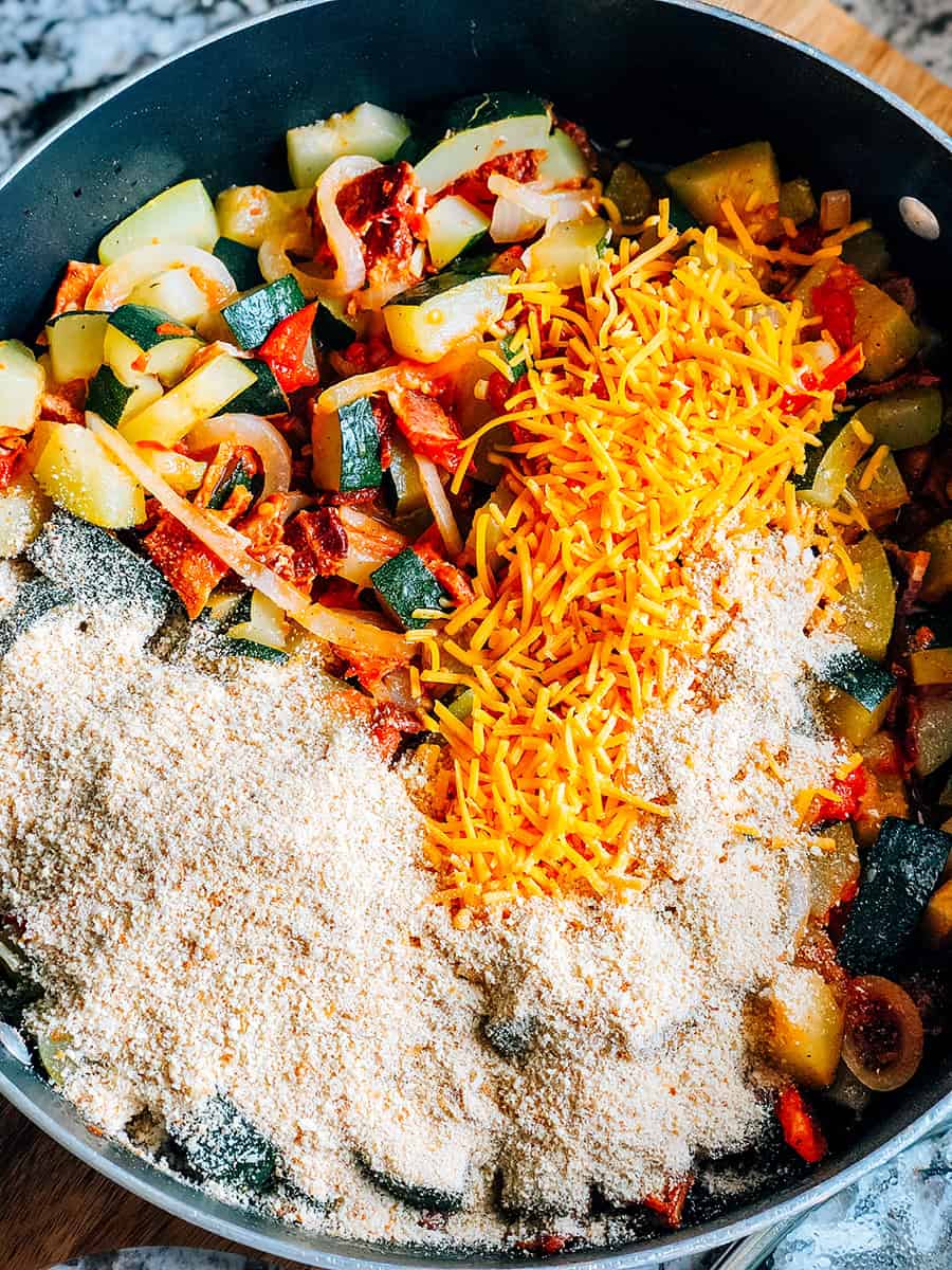 Une poêle pleine de légumes cuits avec du fromage cheddar râpé, de la chapelure, du fromage parmesan râpé et de la sauce soja ajoutée et prête à être mélangée.