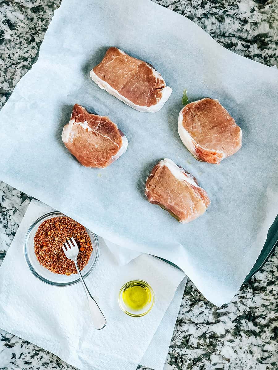 Une vue d'ensemble montrant quatre côtelettes de porc crues sur une plaque de cuisson, un bol de mélange d'épices parfait pour les côtelettes de porc et un peu d'huile d'olive.