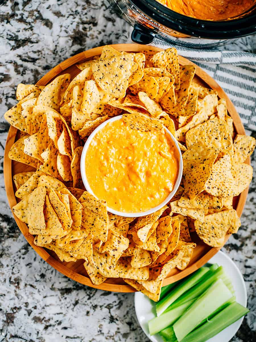 Dip buffalo au poulet crémeux dans un bol de service entouré de chips tortilla épaisses et robustes avec une assiette de bâtonnets de céleri à proximité et la mijoteuse avec du dip supplémentaire dans le coin.