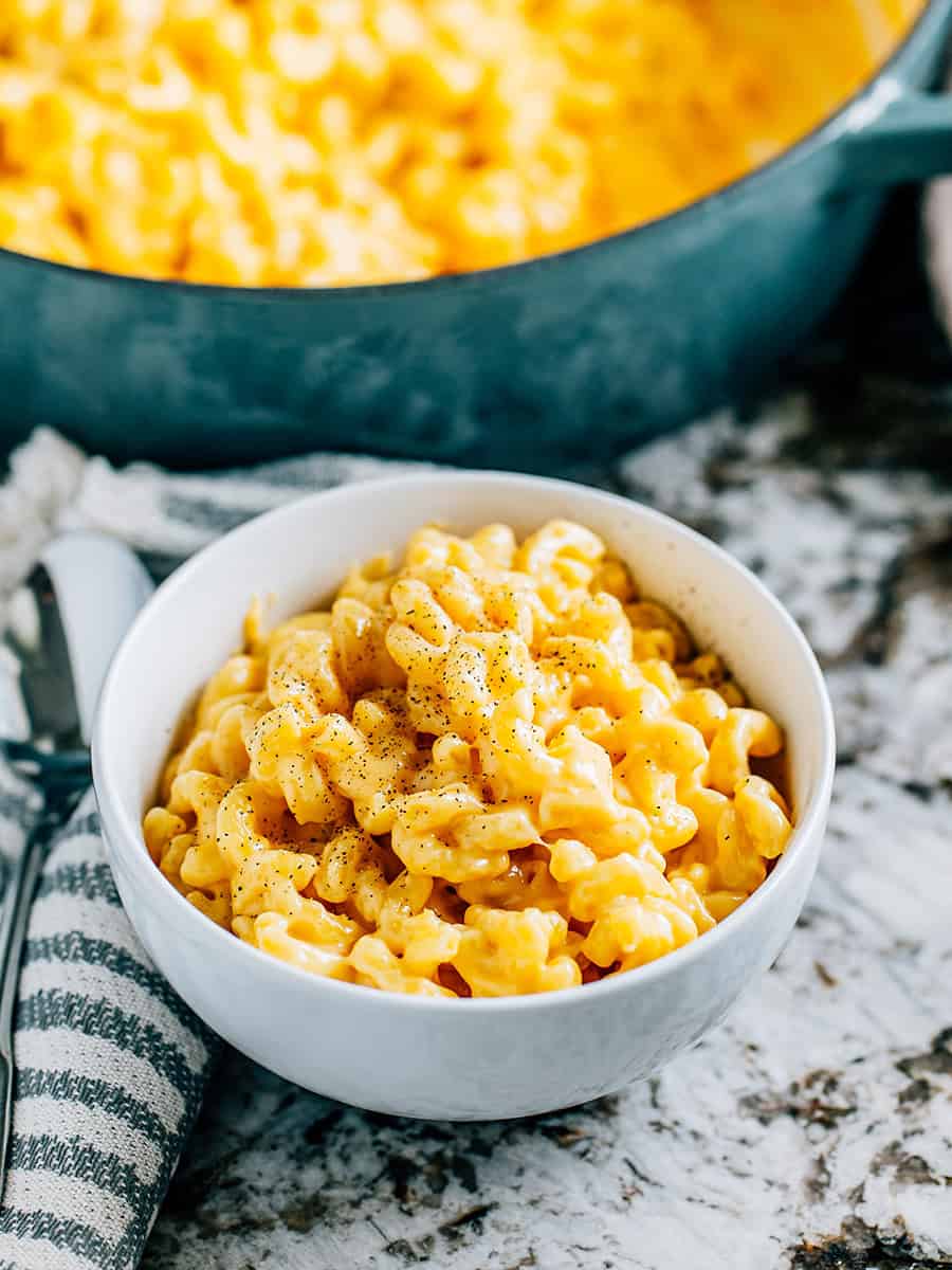 Un bol terminé de macaroni au fromage crémeux fait maison avec une pincée de poivre noir sur le dessus, devant la poêle de macaroni au fromage.