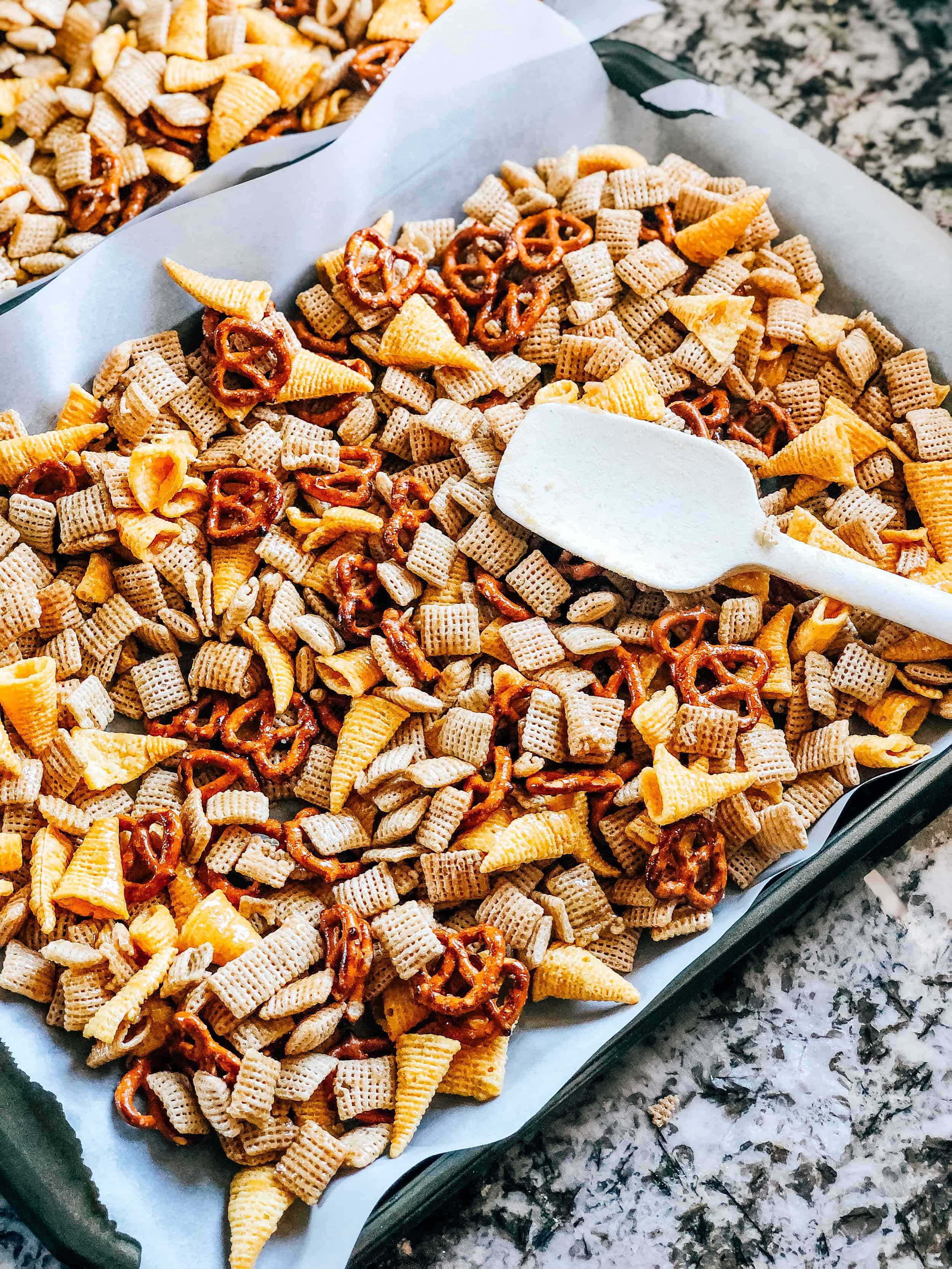 The fall snack mix spread in a thin layer evenly over two large parchment paper lined baking sheets.