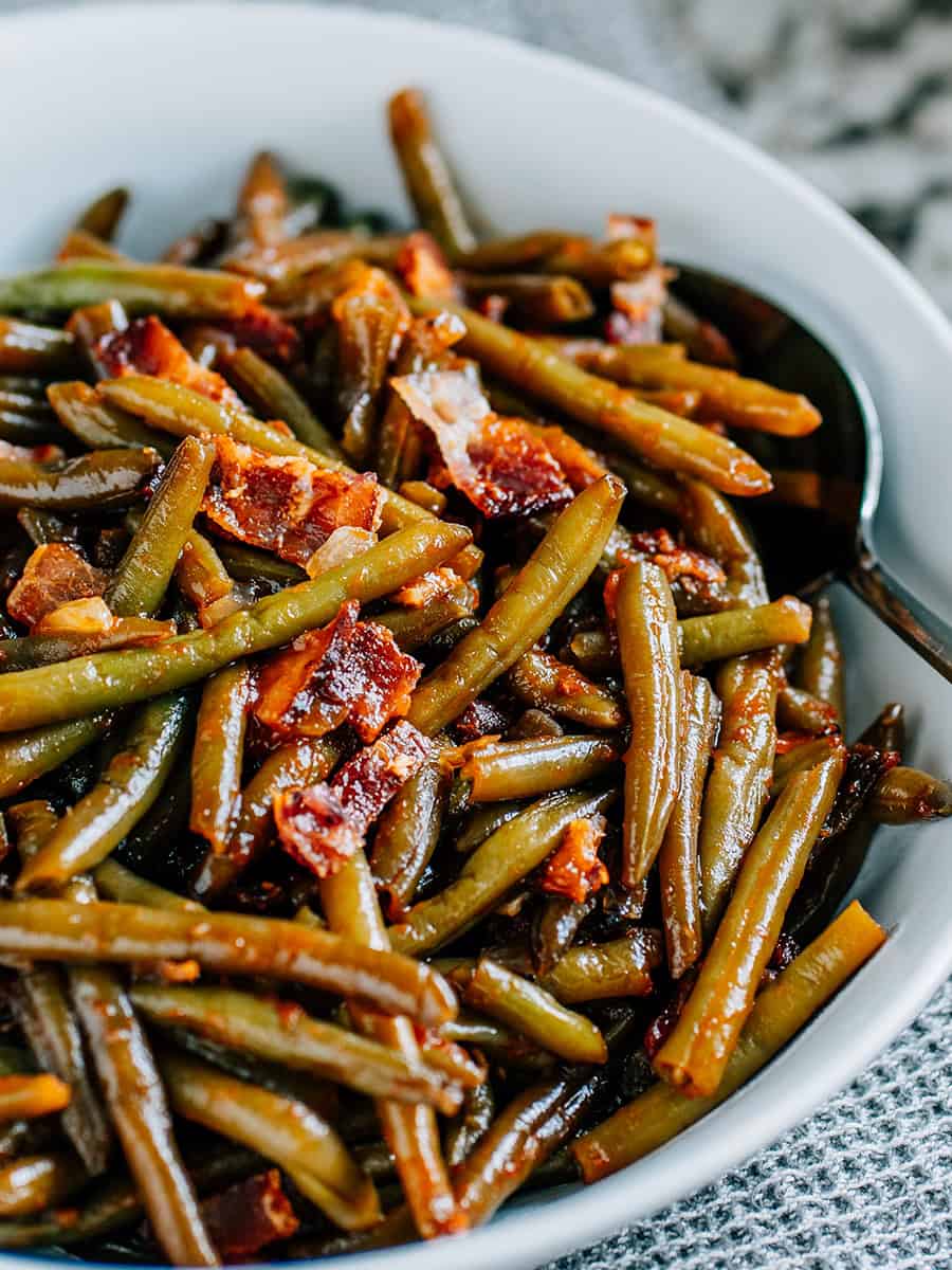 Morceaux de bacon et oignons sur des haricots verts enrobés de BBQ dans un plat de service rond blanc.