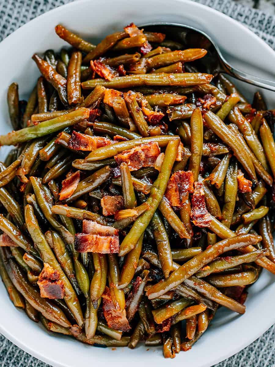 Morceaux de bacon et oignons sur des haricots verts enrobés de BBQ dans un plat de service rond blanc.