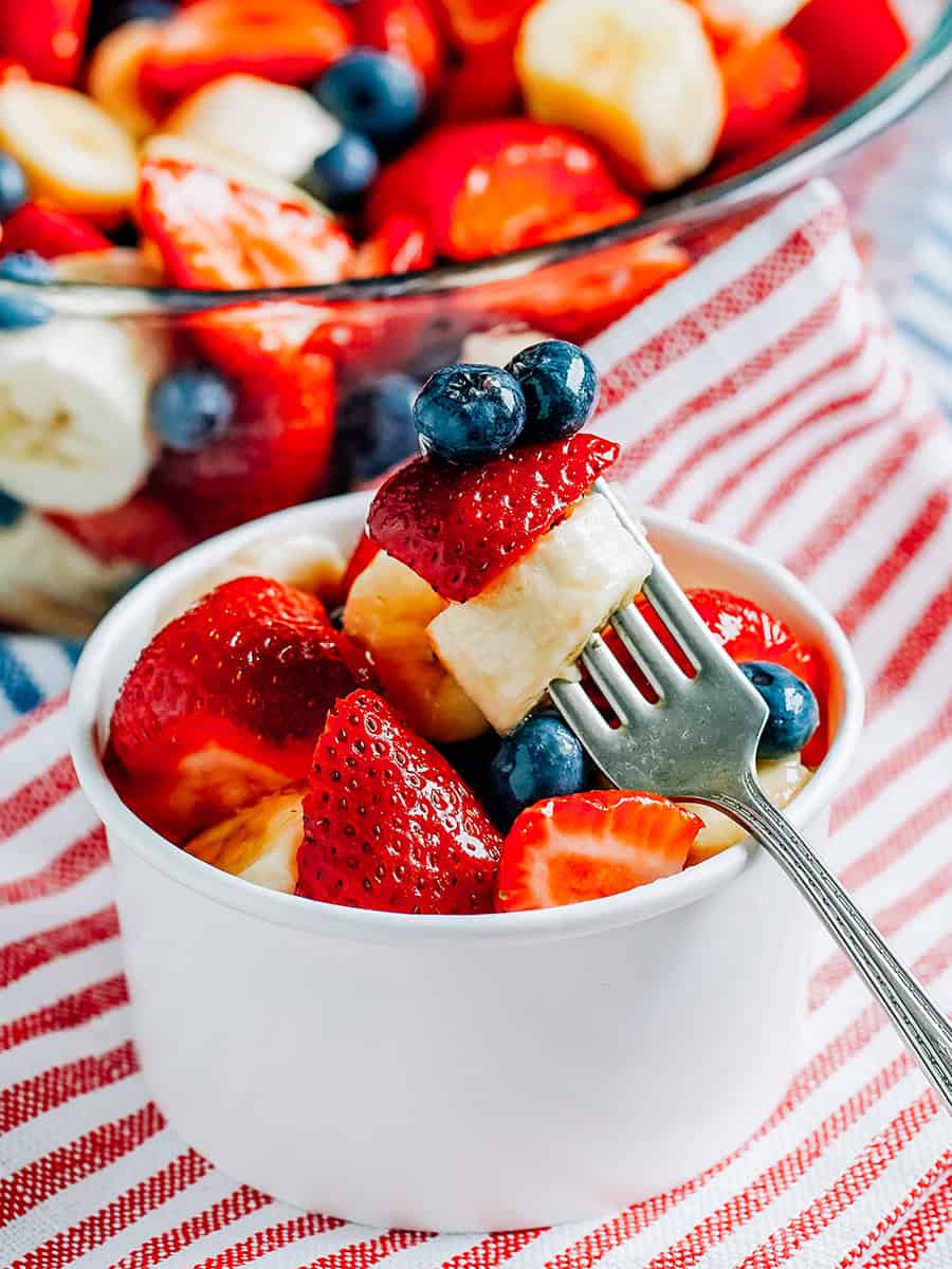 Fraises fraîches, bleuets et bananes enrobés de miel et de lime dans un bol en papier avec des fruits dans un bol de service derrière.