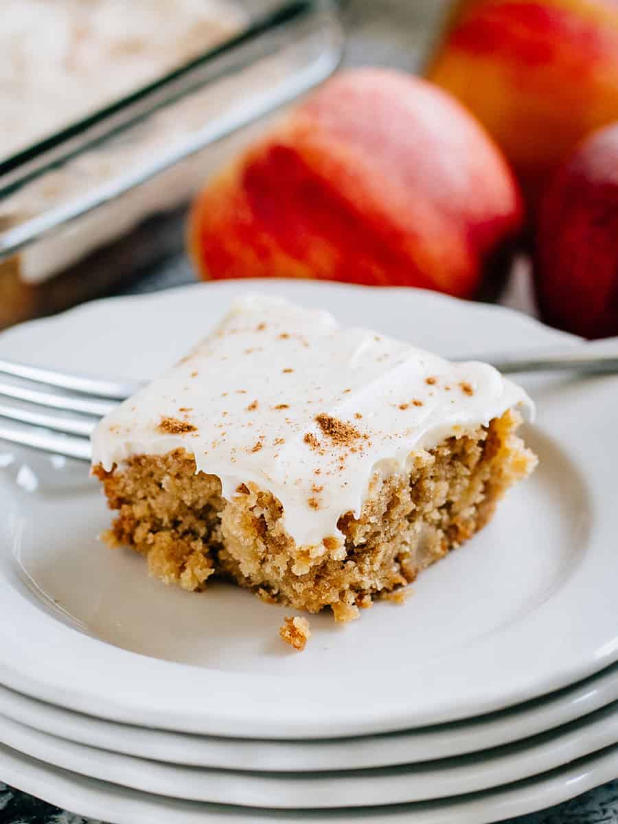 Un blondie aux pommes avec un glaçage au fromage à la crème à l'érable sur une assiette de service avec des pommes en arrière-plan.