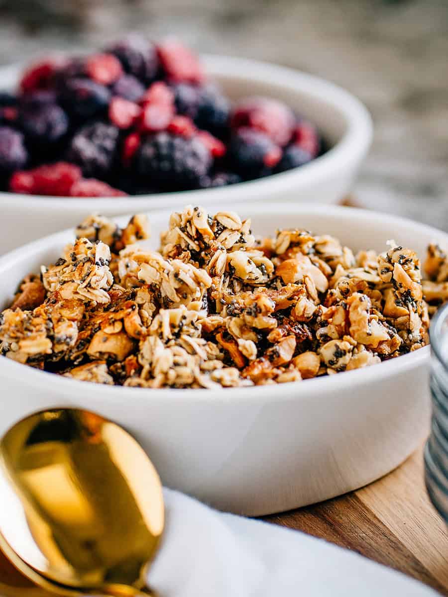 Granola sain fait maison dans un plat de service avec des baies mélangées congelées en arrière-plan, prêt à être servi.