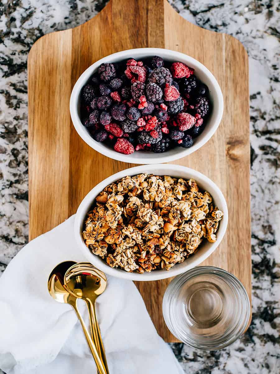 Vue d'un meilleur granola sain fait maison dans un plat de service avec des baies mélangées congelées en arrière-plan, prêt à être servi.