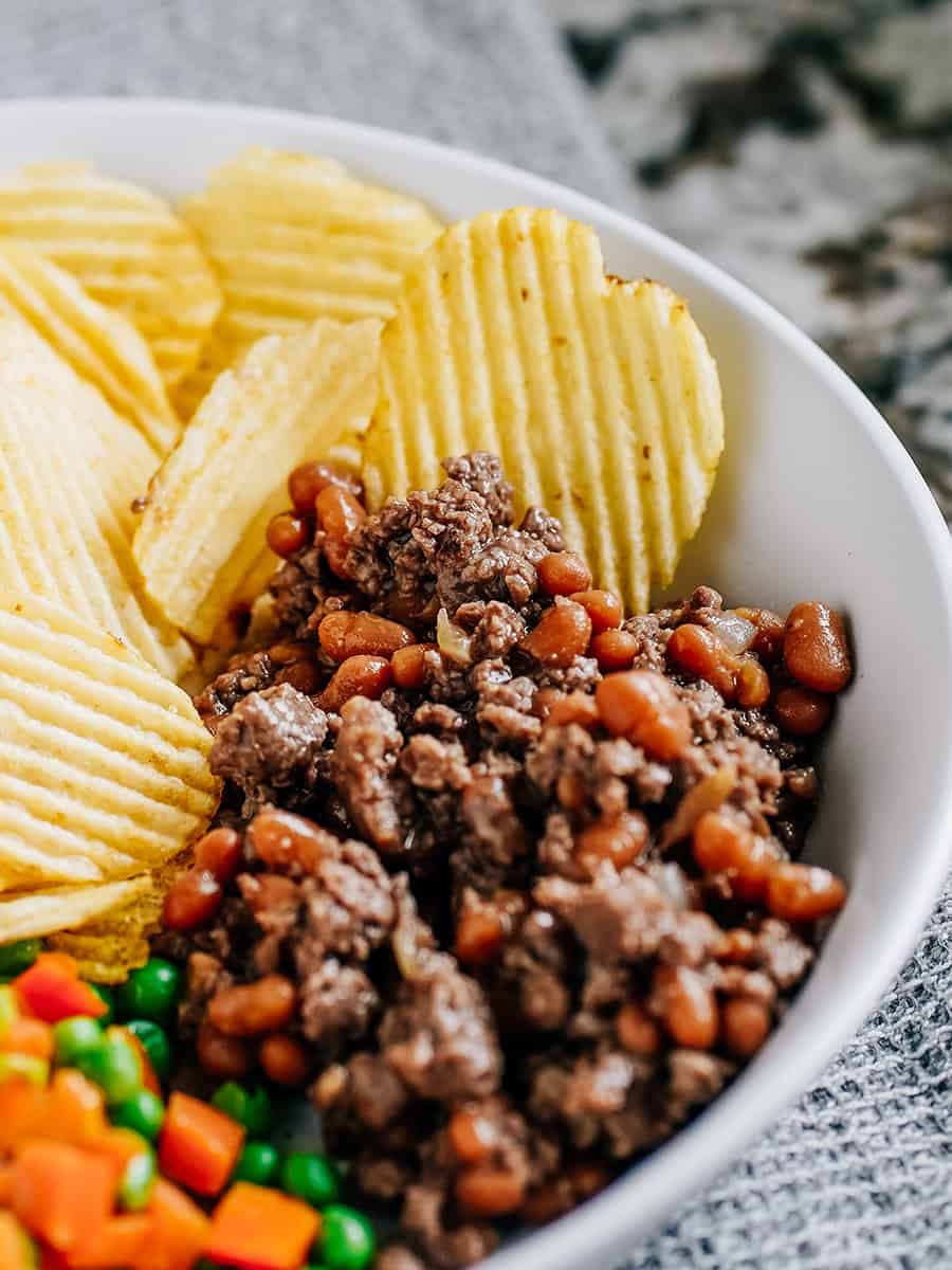 Une assiette de viande hachée et de haricots au four avec des chips et des légumes vapeur à côté.