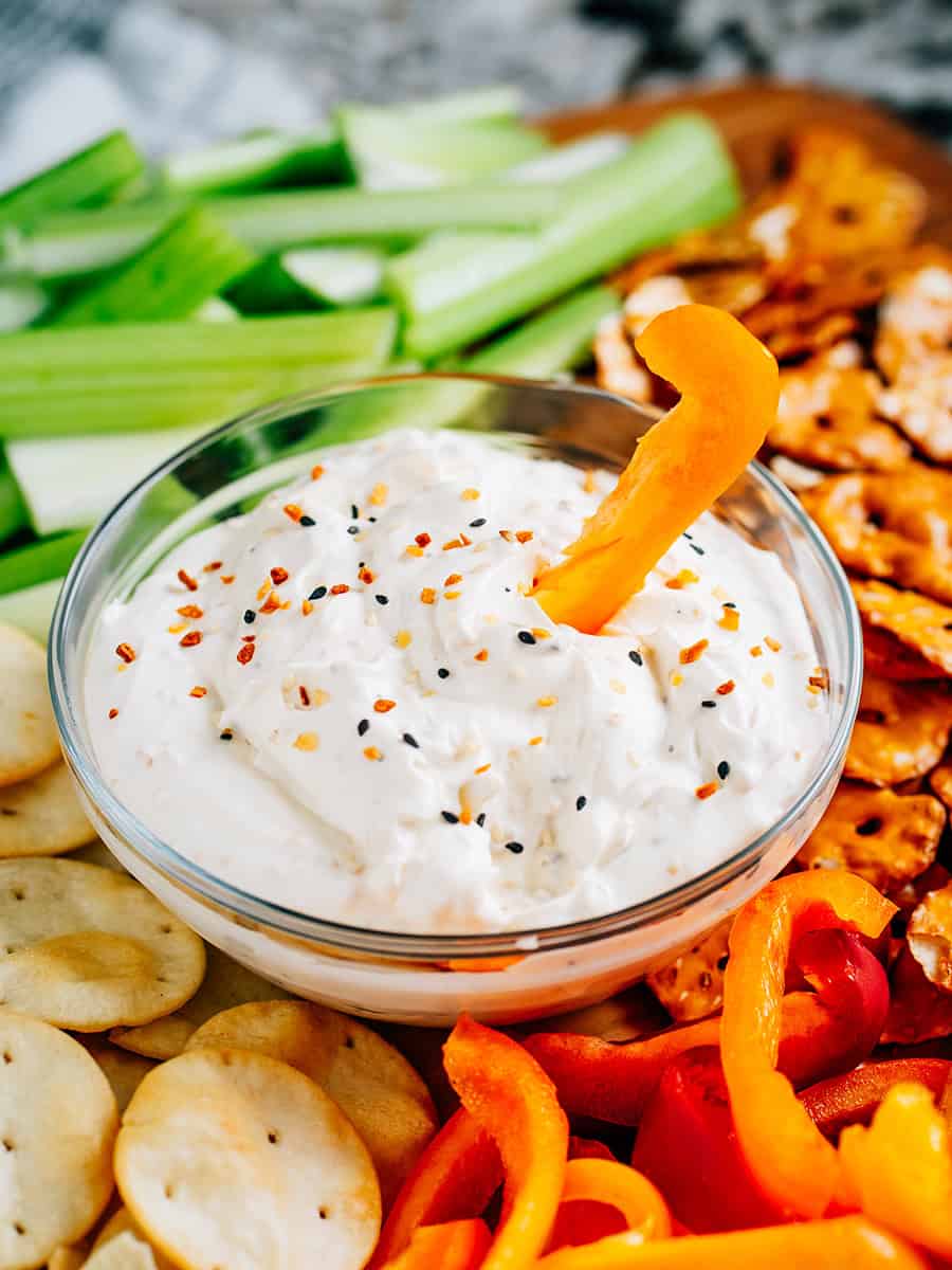 Tartinade de bagel tout en un dans un bol en verre avec un poivron orange à l'intérieur sur un grand plateau en bois entouré de divers dippers.