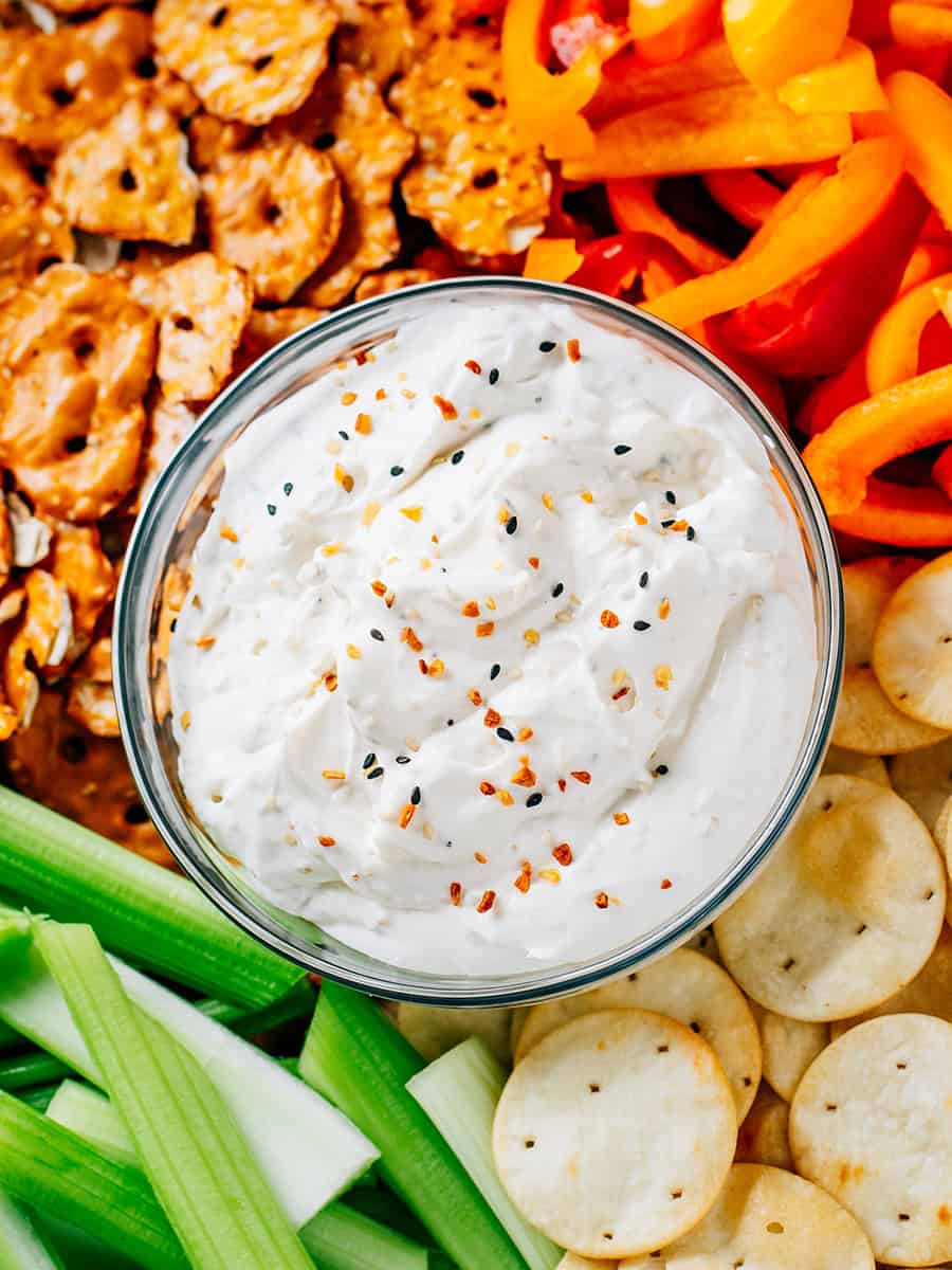 Gros plan de la tartinade de bagel tout en un dans un bol en verre sur un grand plateau en bois entouré de croustillants au bretzel, poivrons, crackers pita et céleri.