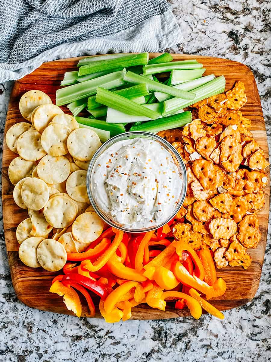 Tartinade de bagel tout en un dans un bol en verre sur un grand plateau en bois entouré de croustillants au bretzel, poivrons, crackers pita et céleri.