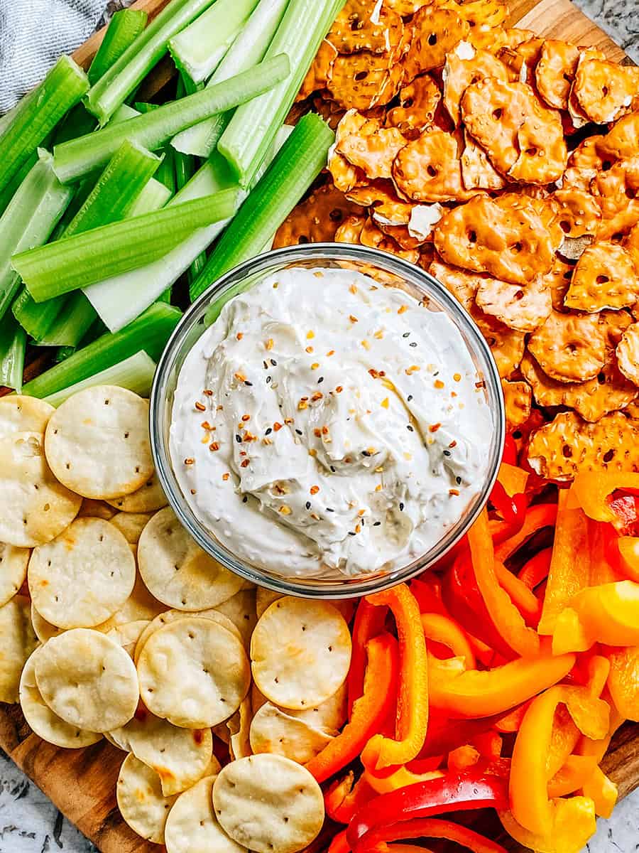 Gros plan de la tartinade de bagel tout en un dans un bol en verre sur un grand plateau en bois entouré de croustillants au bretzel, poivrons, crackers pita et céleri.