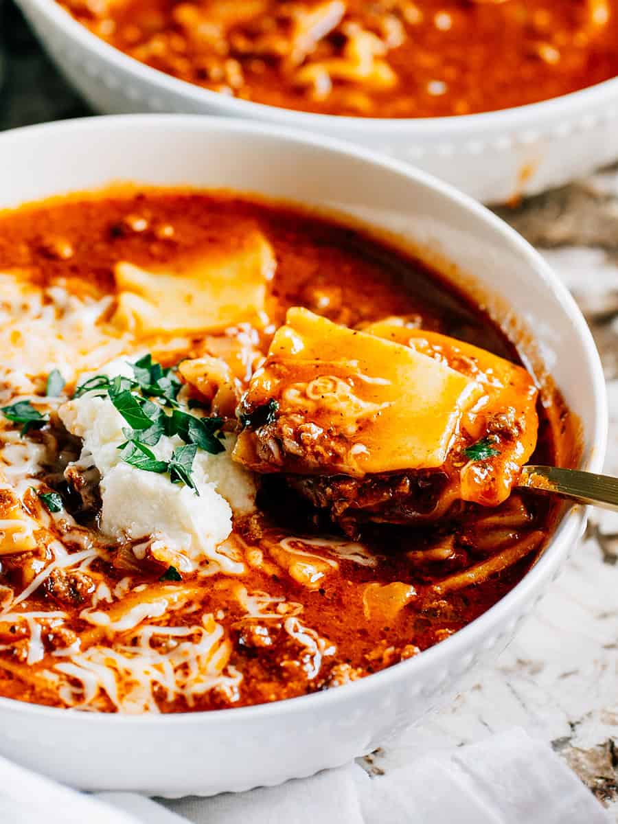 Vue latérale d'une cuillère de soupe en train d'être soulevée d'un bol de soupe blanc. Un bol de soupe de lasagne chaude et réconfortante avec du boeuf dans un bouillon de boeuf et marinara, garni d'une cuillerée de ricotta, de fromage cheddar râpé et de persil.