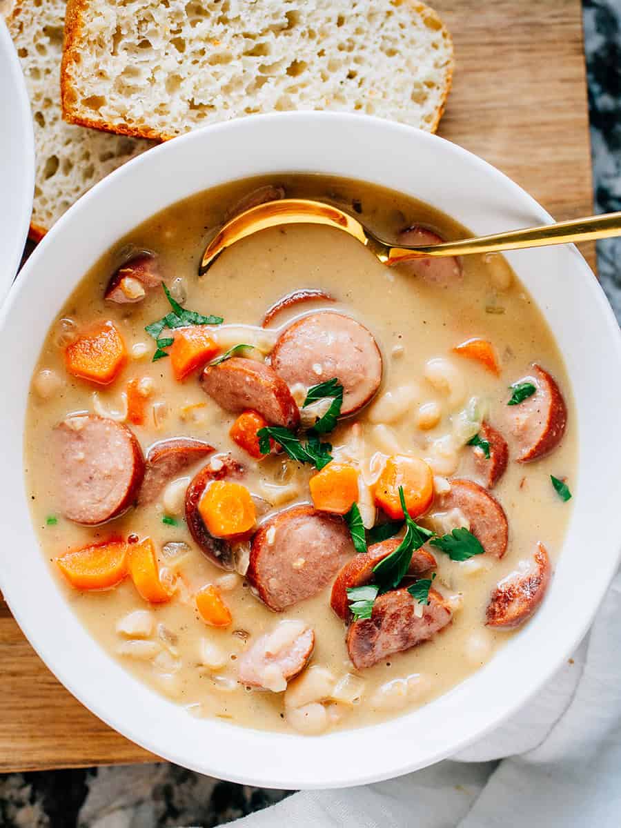 Vue de dessus : Un bol de kielbasa savoureuse, de carottes cuites tendres et de haricots crémeux Great Northern mijotés à perfection dans une sauce crémeuse avec de l'ail haché et des oignons. C'est la meilleure soupe aux haricots ! Notre célèbre pain paysan fait maison en accompagnement.