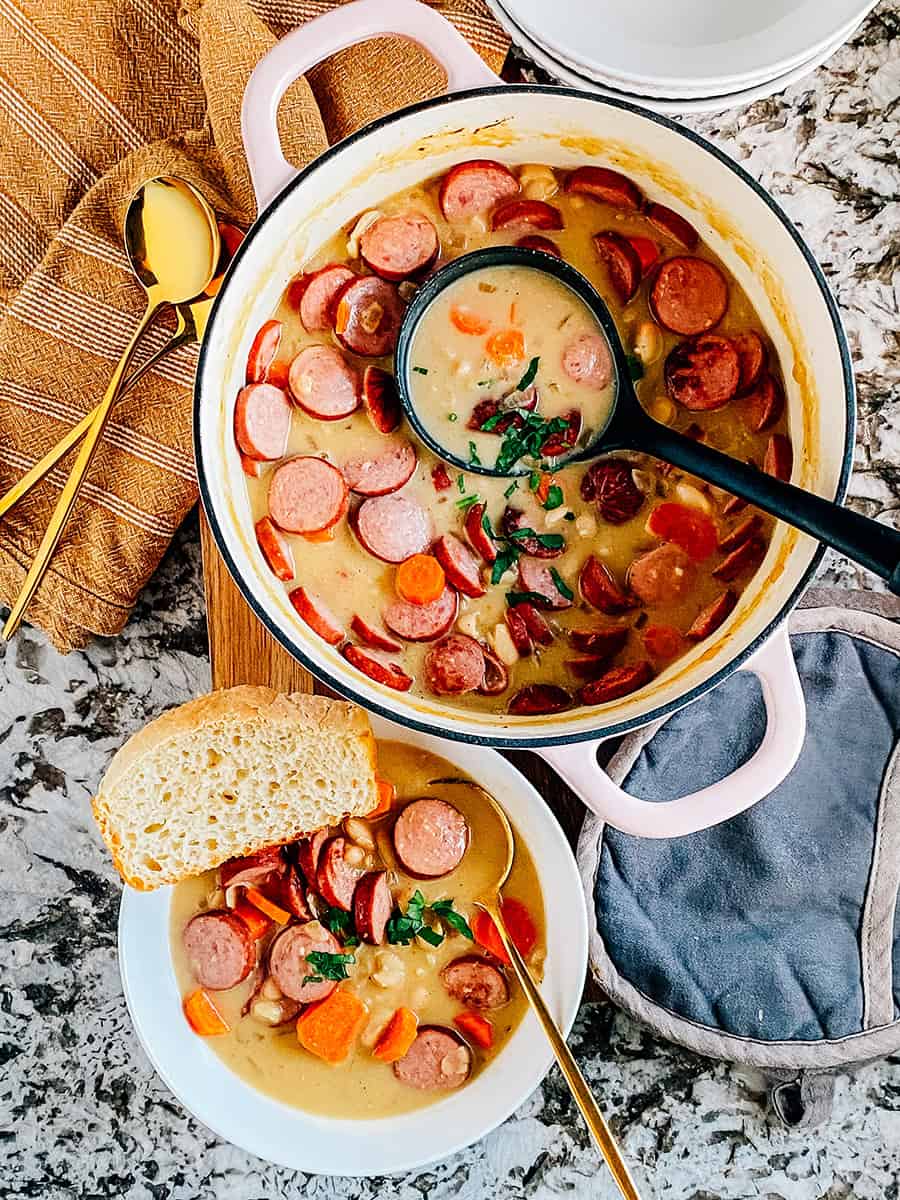 Une casserole pleine de morceaux de kielbasa dorés, de carottes cuites tendres et de haricots crémeux Great Northern mijotés à perfection dans une sauce crémeuse avec de l'ail haché et des oignons. C'est un plat savoureux plein de saveurs. Un bol de soupe dans le coin inférieur gauche avec du pain fraîchement cuit à côté.