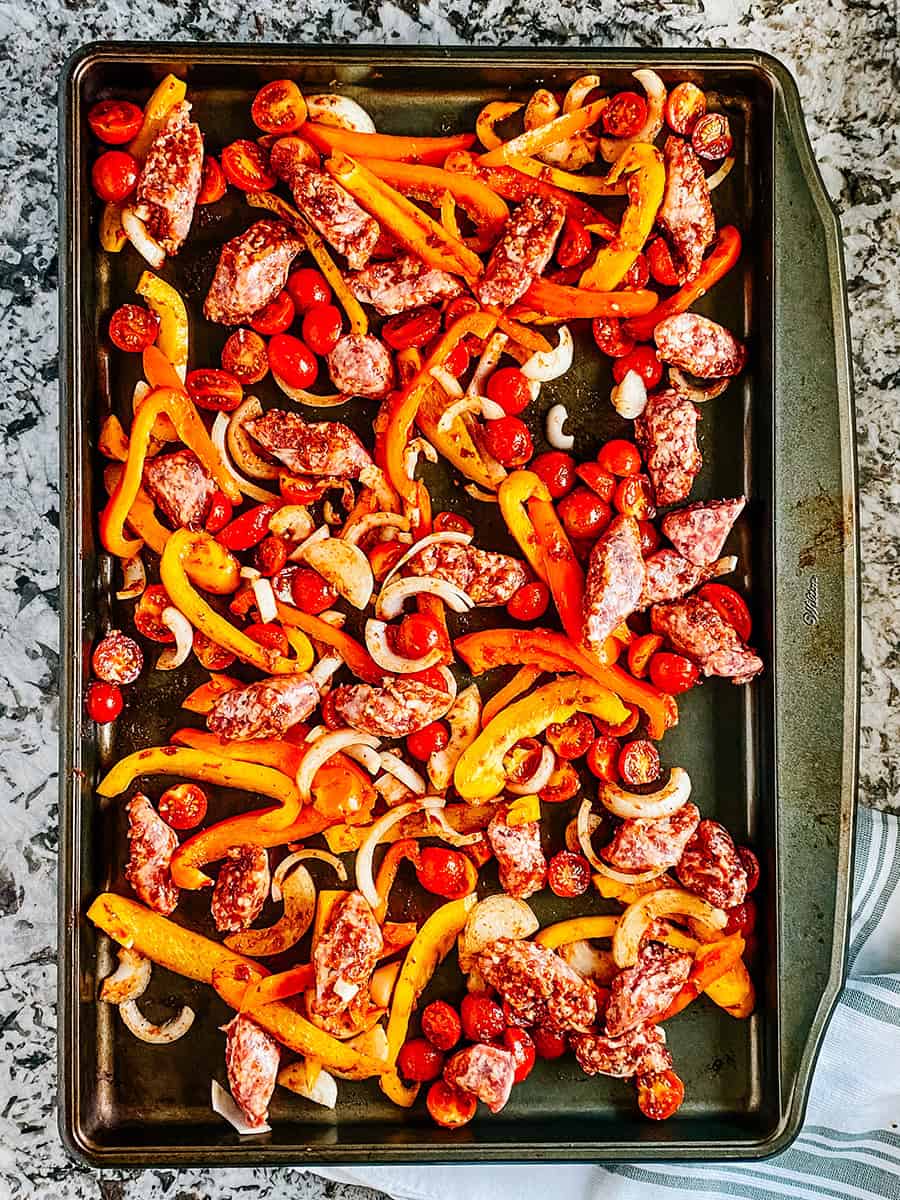 Uncooked sausage and sliced vegetables coated in a balsamic vinegar homemade marinade spread out on a rimmed baking sheet.