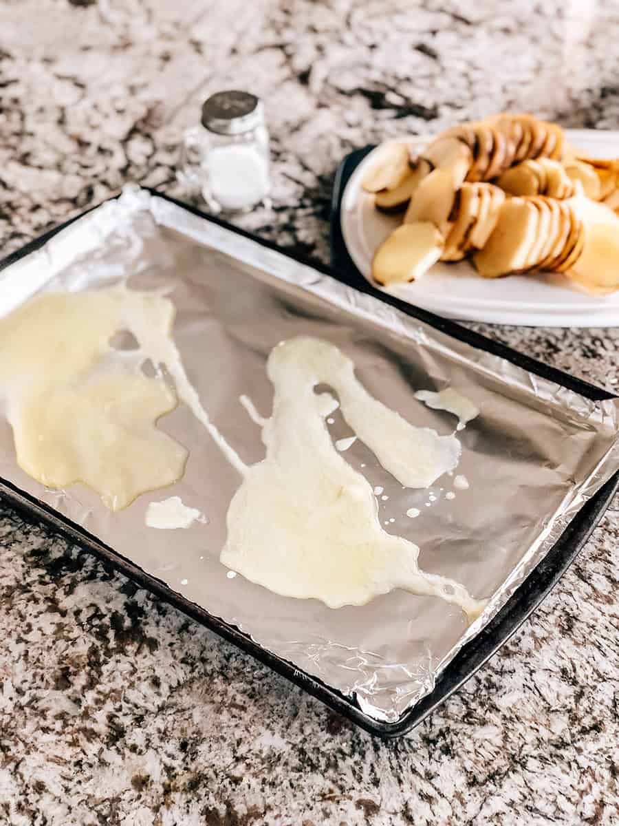 Plaque de cuisson tapissée de papier aluminium et arrosée de beurre fondu avec des tranches de pommes de terre en arrière-plan sur une planche à découper.