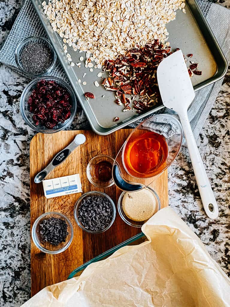 Ingrédients pour la meilleure recette de barre granola moelleuse : flocons d'avoine, noix, fruits secs, graines de chia, miel, beurre, sel, vanille, sucre brun et pépites de chocolat !